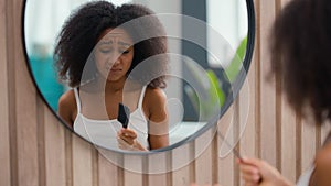 Frustrated African American woman suffer problem trouble curls looking at mirror at bathroom beauty routine prepare