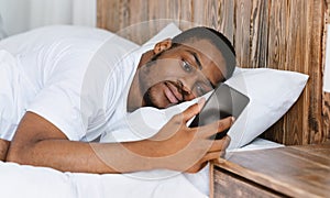 Frustrated African Guy Looking At Mobile Phone Lying In Bed