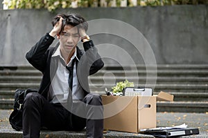 A frustrated Asian businessman sits on the stairs with a box after getting fired