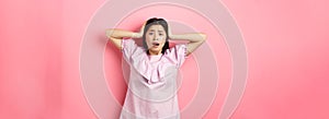 Frustrated asian teen girl panicking, holding hands on head and screaming scared, standing anxious on pink background