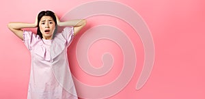 Frustrated asian teen girl panicking, holding hands on head and screaming scared, standing anxious on pink background