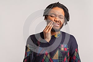 Frustrated bearded african man with dreadlocks touching cheek and wincing in pain feeling terrible tooth ache, gingivitis,