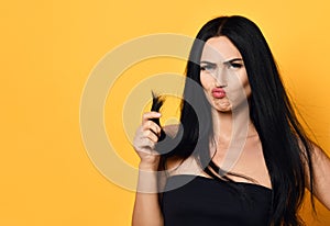 Frustrated beautiful brunette woman examining looking at her dry damaged hair over yellow background. Health And Beauty Concept