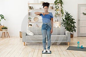 Frustrated Black Lady Gesturing Thumbs-Down Standing On Scales Indoors