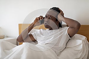 Frustrated Black Man Talking On Cellphone While Sitting In Bed At Home
