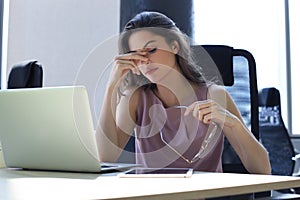 Frustrated business woman looking exhausted while sitting at her working place and carrying her glasses in hand