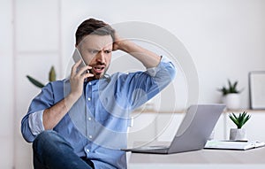 Frustrated businessman having problems with computer, talking on cellphone with customer support