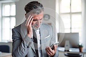 A frustrated businessman with smartphone standing in an office, reading bad news.