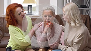 Frustrated desperate Caucasian woman thinking sitting on couch in living room as empathetic friends calming down