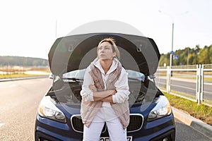 frustrated female driver sits by a broken down car on the hood waiting for help, transport auto insurance concept