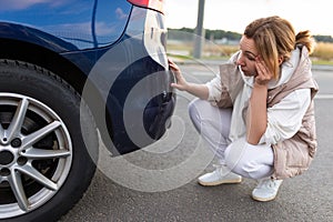 frustrated female driver sits at the rear bumper of the car and examines the damage and scratches, traffic accident