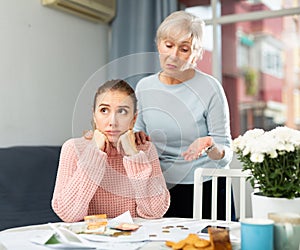 Frustrated girl and elderly woman counting cash at table at home