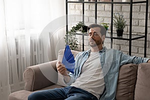 Frustrated guy recline on sofa cool down with hand fan