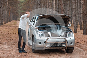 A frustrated guy, standing near a broken car with an open hood with smoke, stands and holds head.