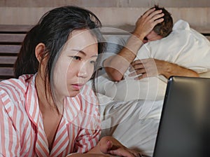Frustrated husband moody in bed ignored by his workaholic Asian wife or internet social media addict girlfriend using laptop in