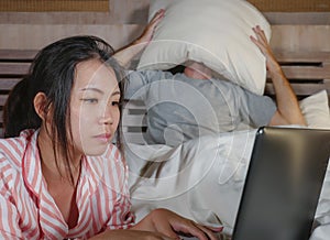 Frustrated husband moody in bed ignored by his workaholic Asian wife or internet social media addict girlfriend using laptop in