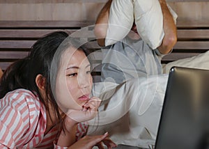 Frustrated husband moody in bed ignored by his workaholic Asian wife or internet social media addict girlfriend using laptop in