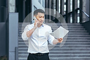 Frustrated insurance agent with documents talking on the phone outside the office, depressed Asian businessman
