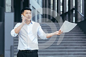 Frustrated insurance agent with documents talking on the phone outside the office, depressed Asian businessman