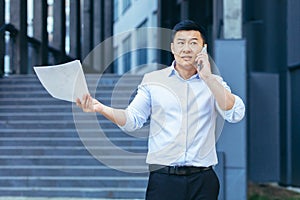 Frustrated insurance agent with documents talking on the phone outside the office, depressed Asian businessman