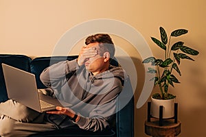 Frustrated man lying on the couch with laptop. Covering face with his hand