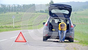 Frustrated man sitting in open trunk of broken car on empty countryside road