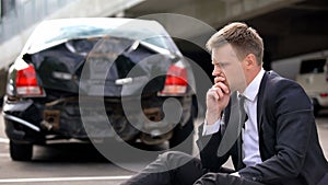 Frustrated man sitting road on crashed car background, traffic accident stress