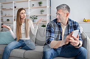 Frustrated mature woman arguing with her husband about smartphone overuse, expressing jealousy at home