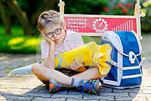 Frustrated sad little kid boy with glasses sitting by desk and backpack or satchel. Schoolkid with traditional German