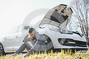 Frustrated sad upset guy driver is sitting near a broken car after road accident, crash. Young shocked desperate