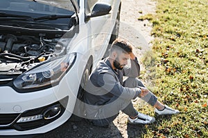 Frustrated sad upset guy driver is sitting near a broken car after road accident, crash. Young shocked desperate