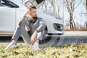 Frustrated sad upset guy driver is sitting near a broken car after road accident, crash. Young shocked desperate