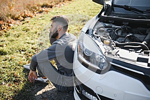 Frustrated sad upset guy driver is sitting near a broken car after road accident, crash. Young shocked desperate