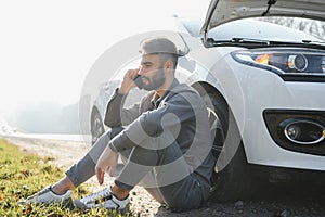 Frustrated sad upset guy driver is sitting near a broken car after road accident, crash. Young shocked desperate