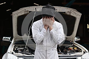 Frustrated stressed young mechanic man in white uniform covering his face with hands against car in open hood at the repair garage