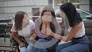 Frustrated teenage girl crying as friends calming down sitting on bench on city street. Portrait of troubled depressed