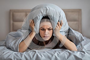 Frustrated woman covering ears with pillow, annoyed by noise