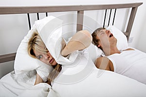 Frustrated woman covering ears with pillow while man snoring in bed