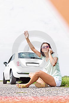 Frustrated woman hitchhiking while using cell phone on country road by broken down car