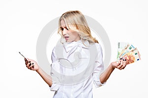 Frustrated woman looking in disbelief at her mobile phone, holding loads of Euro banknotes against white background. Phone bill.