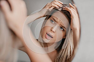 Frustrated woman searching hair flakes suffering from dandruff problem, looking at her reflection in mirror in bathroom