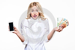 Frustrated woman shrugging shoulders, showing mobile phone blank screen and holding loads of Euro banknotes on white background. photo