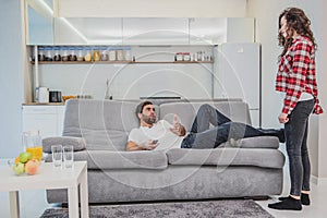 A frustrated woman talking to her husband while watching a TV. A man sitting on the couch shows a gesture with his