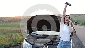 A frustrated young beautiful girl is standing by a broken car on the highway at sunset and calls the car service