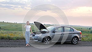 A frustrated young beautiful girl is standing by a broken car on the highway at sunset and calls the car service. An