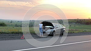 A frustrated young beautiful girl is standing by a broken car on the highway at sunset and calls the car service. An