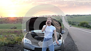 A frustrated young beautiful girl is standing by a broken car on the highway at sunset and calls the car service