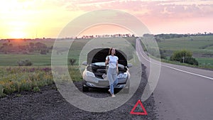 A frustrated young beautiful girl is standing by a broken car on the highway at sunset and calls the car service. An