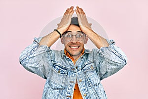 A frustrated young man dressed in a denim jacket and transparent spectacles keeping both hands on his head isolated on pink