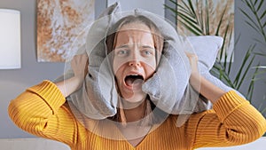 Frustrated young screaming woman covering ears with pillow feeling panic suffering from loud noise keeps mouth opened looks scared